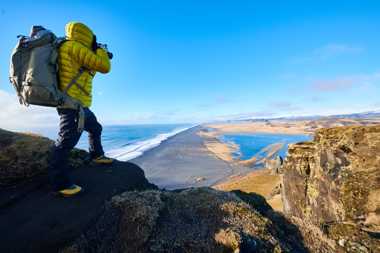 Male wearing a yellow jacket standing on a rock while taking a picture of the beautiful scenery